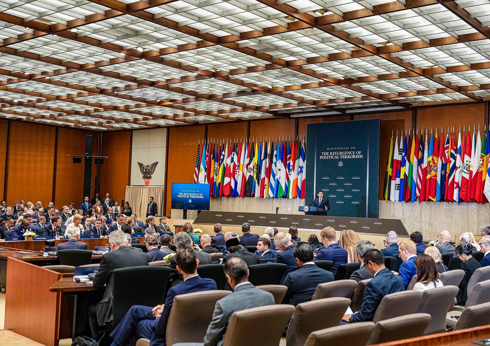 Delegates seated in a State Department hall lined with national flags as Secretary Marco Rubio opens the Ministerial on the Resurgence of Political Terrorism, July 16, 2026.
