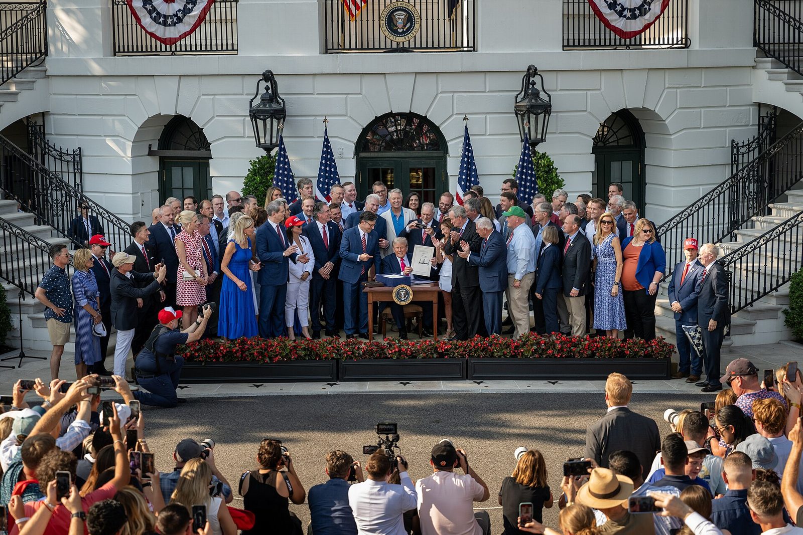 President Trump signs the One Big Beautiful Bill Act at a table on the South Lawn of the White House, surrounded by lawmakers and officials, with press photographers in the foreground.