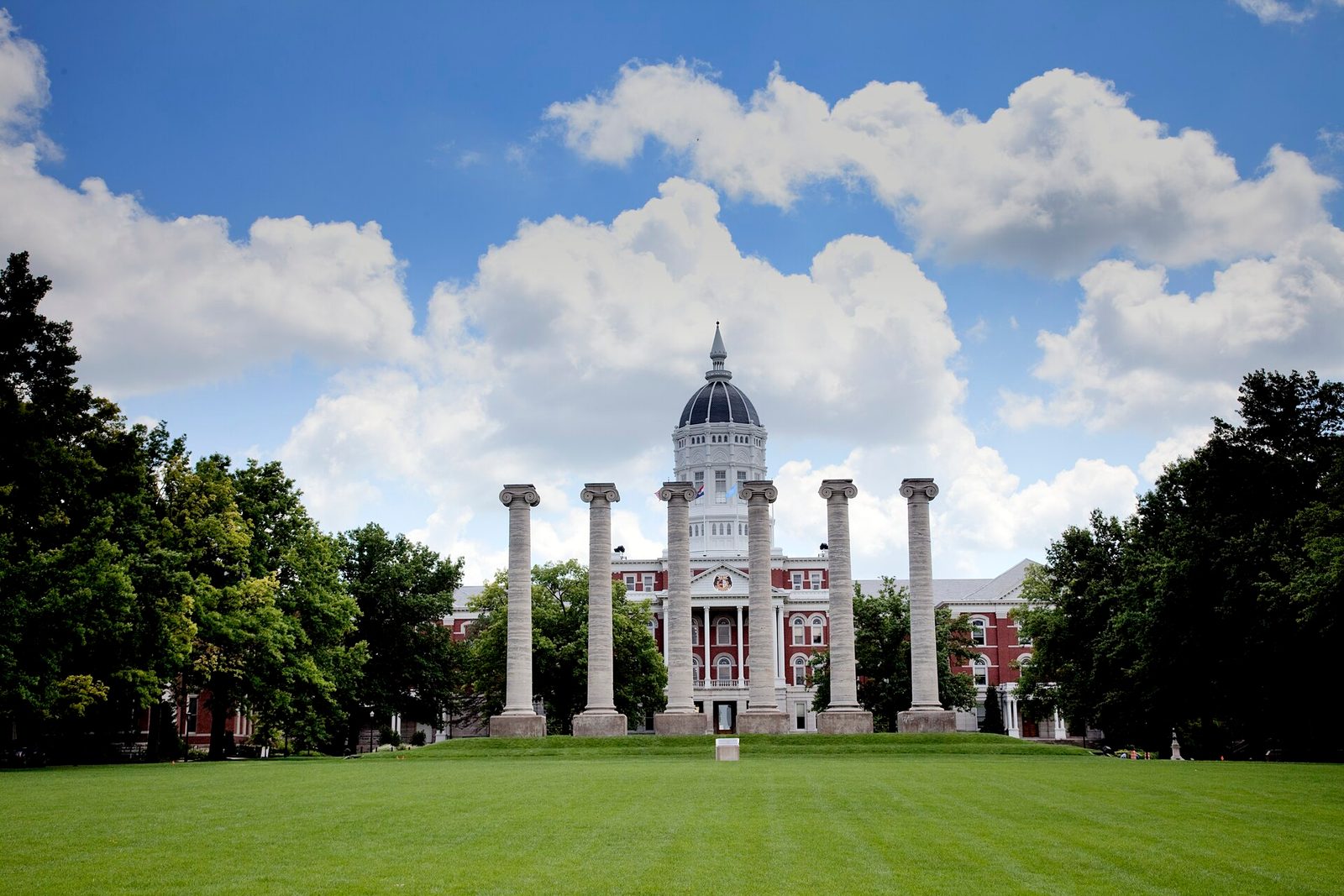 The six stone columns of the Francis Quadrangle in front of the domed Jesse Hall at the University of Missouri, under a cloudy blue sky.