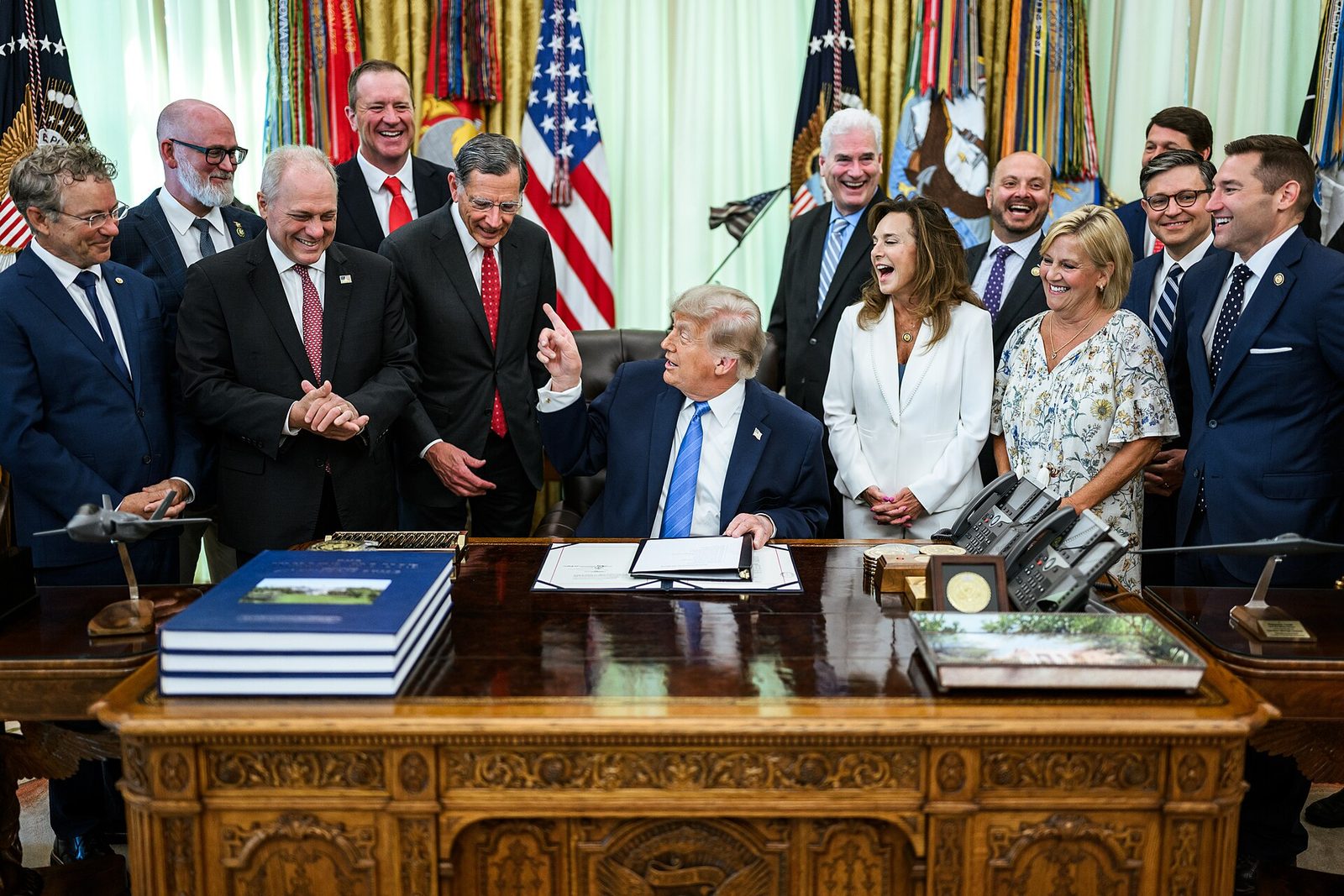 President Trump sits at the Resolute Desk in the Oval Office with lawmakers standing behind him at the signing of the Secure America Act.