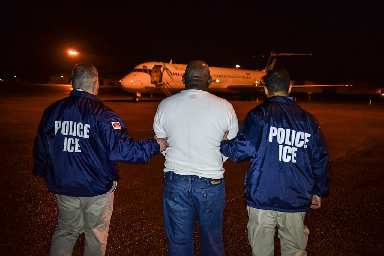 ICE officers escort a detainee across an airfield toward a waiting ICE Air charter aircraft.
