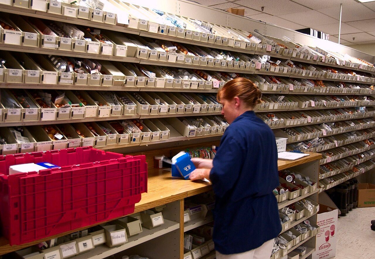 A hospital pharmacist in scrubs restocks medication into rows of labeled bins at a Navy medical center pharmacy.